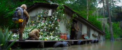 a small house on a river with a mass of ivy and flower crawling up one side. josie is kneeling at the foot, looking at the flowers, while ventress supervises